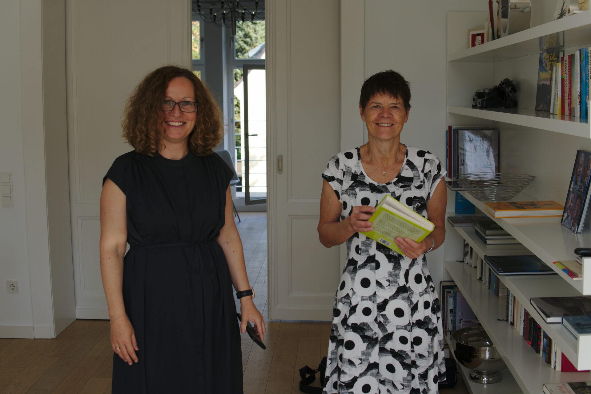 Katja Schumacher, eine Frau mit schulterlangen Locken und freundlichem Lächeln steht neben Angela Lechner, eine Frau mit kurzen dunklen Haaren, offenem Lächeln und einem Buch in der Hand. Neben ihnen steht ein Bücherregal. Beide Frauen tragen Kleider.