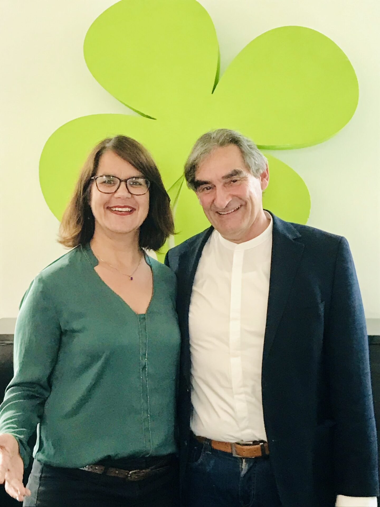 Peter Schönfelder und Esther Himmen stehen bei einem Jobsharing-Coaching vor einem grünen, vierblättrigen Kleeblatt. Sie sind formell gekleidet.